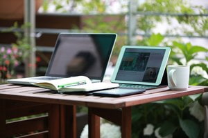 Schreibtisch mit Laptop, Tablet, Smartphone, einem Notizbock und einer Kaffeetasse. Pflanzen im Hintergrund